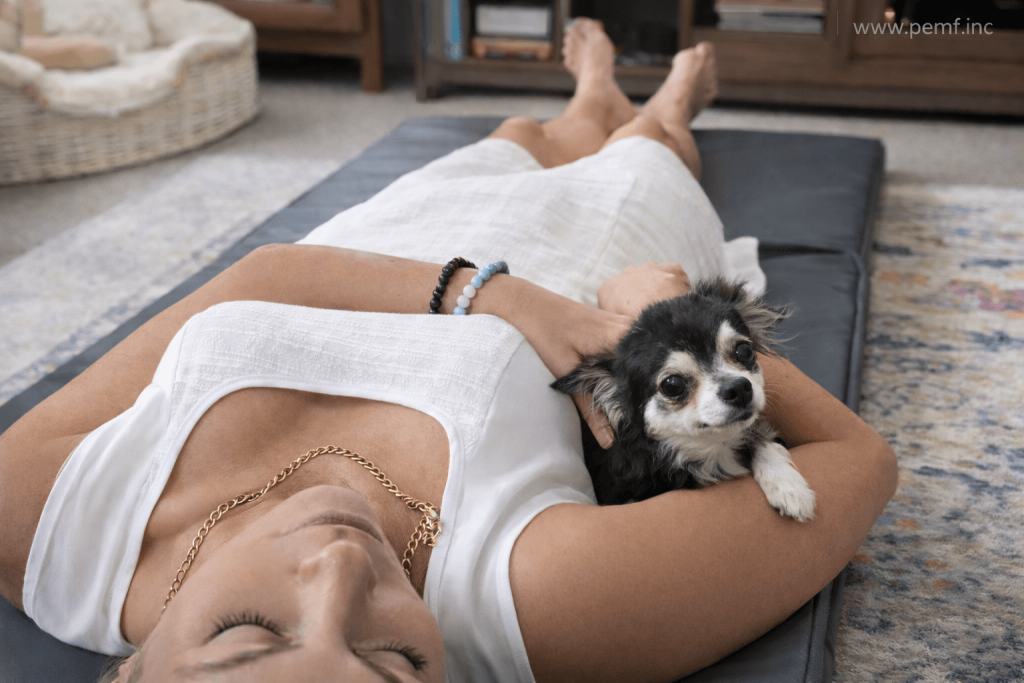 Woman relaxing on a PEMF mat at home with a small dog resting beside her